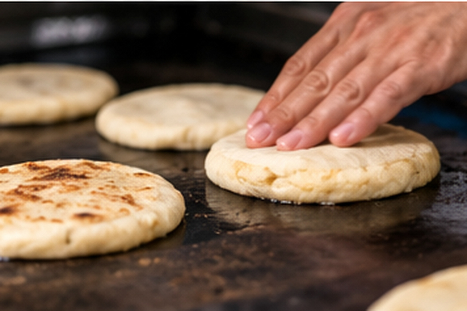 Una mano forma una pupusa gruesa sobre el comal mientras otras se doran.