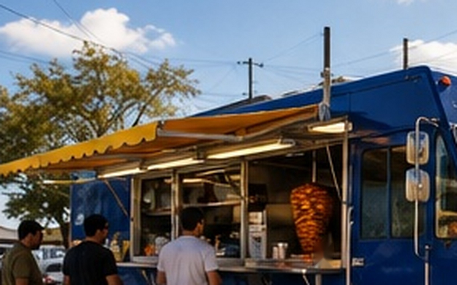 Tres clientes esperan frente a la ventanilla abierta del taco truck, con el trompo visible en el interior.